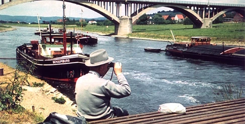 belebete-Weser-frueher.jpg Ein Mann mit Fernglas beobachtet Schiffe auf der Weser, während eine beeindruckende Brücke im Hintergrund das Flussufer überquert.
