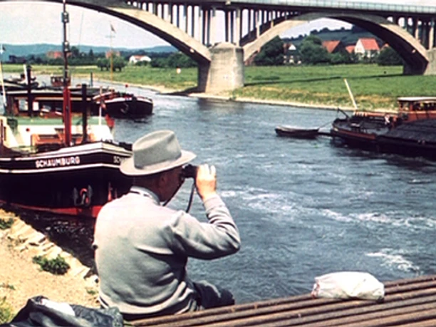 belebete-Weser-frueher.jpg Ein Mann mit Fernglas beobachtet Schiffe auf der Weser, während eine beeindruckende Brücke im Hintergrund das Flussufer überquert.