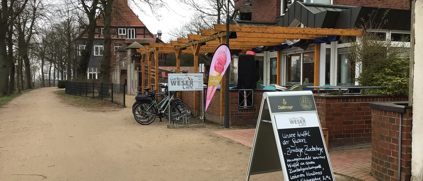 A path leads along the terrace of Garbe's Weser Café, surrounded by trees and a building.