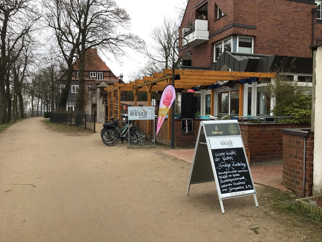 Garbes Weser-Café Ein Weg führt an der Terrasse von Garbes Weser-Café entlang, umgeben von Bäumen und einem Gebäude.