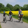 Drei Radfahrer, darunter ein Kind, fahren einen schmalen Weg entlang, gesäumt von gelben Rapsfeldern.