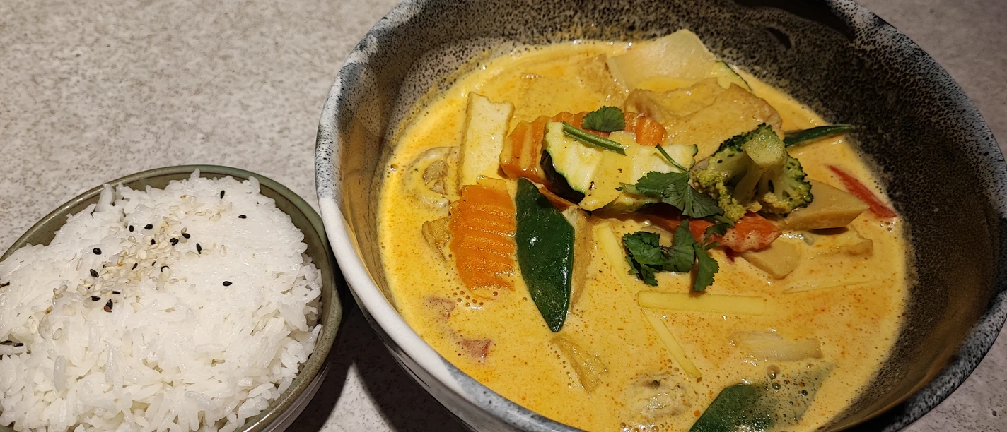 Hähnchen Mango Curry.jpg Eine Schale mit goldgelbem, cremigem Hähnchen-Mango-Curry und bunter Gemüseauswahl. Daneben schneeweißer Reis.