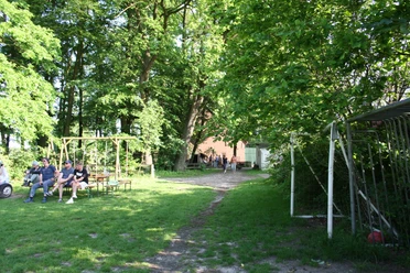 IMG_8639.JPG Grüne Wiese mit Fußballtor, Bierzeltgarnituren und Menschen im Schatten von Bäumen.