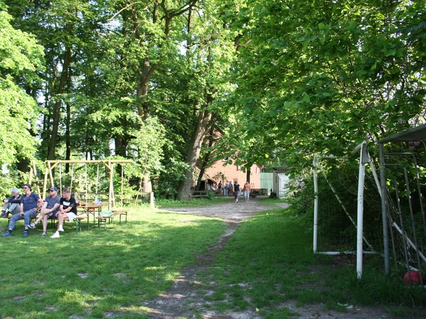 IMG_8639.JPG Grüne Wiese mit Fußballtor, Bierzeltgarnituren und Menschen im Schatten von Bäumen.