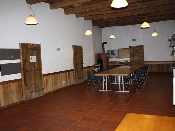 IMG_7950.JPG Ein großer Raum mit Holzbalkendecke, Backsteinofen, langen Tischreihen und Stühlen in warmer Beleuchtung.