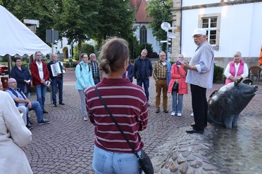 VIP_Fuehrung (4).JPG Eine kleine Menschenmenge hört interessiert einem Mann in historischer Kleidung zu, der auf einem Brunnen steht.