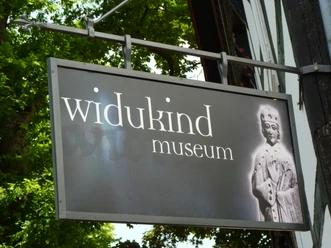 P1090657.JPG Schild des Widukind Museums mit historischer Abbildung eines mittelalterlichen Königs auf schwarzem Hintergrund.