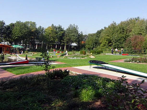 Grüner Minigolfplatz mit mehreren Bahnen, umgeben von Bäumen; Menschen spielen bei sonnigem Wetter.