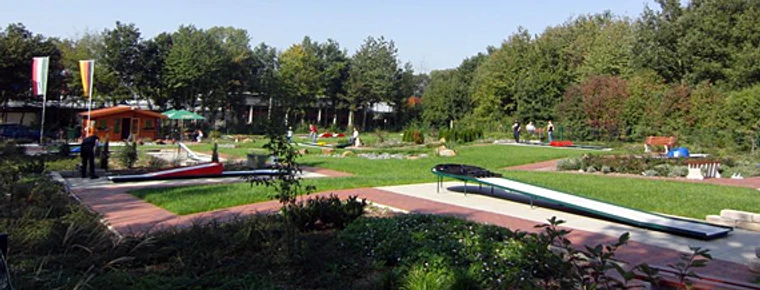 Grüner Minigolfplatz mit mehreren Bahnen, umgeben von Bäumen; Menschen spielen bei sonnigem Wetter.