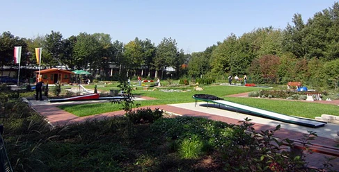 minigolfplatz.jpg Grüner Minigolfplatz mit mehreren Bahnen, umgeben von Bäumen; Menschen spielen bei sonnigem Wetter.