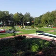 minigolfplatz.jpg Grüner Minigolfplatz mit mehreren Bahnen, umgeben von Bäumen; Menschen spielen bei sonnigem Wetter.