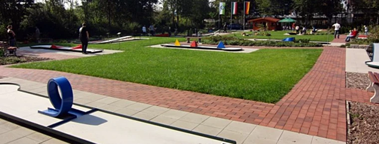 Eine Minigolfanlage mit gepflegtem Rasen, bunten Hindernissen und teils schattigen Plätzen im Freien.