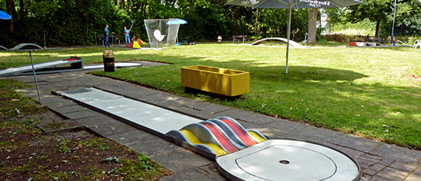Eine Minigolfbahn umgeben von Grünflächen, mit bunten Hindernissen und Sonnenschirmen ausgestattet.
