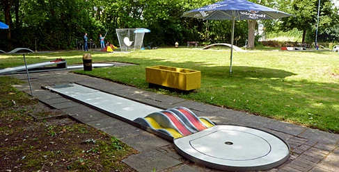 030_minigolf.jpg Eine Minigolfbahn umgeben von Grünflächen, mit bunten Hindernissen und Sonnenschirmen ausgestattet.