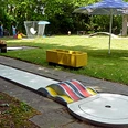 030_minigolf.jpg Eine Minigolfbahn umgeben von Grünflächen, mit bunten Hindernissen und Sonnenschirmen ausgestattet.