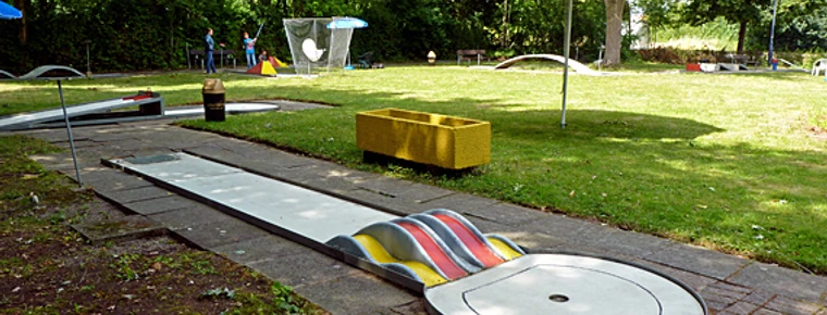 030_minigolf.jpg Eine Minigolfbahn umgeben von Grünflächen, mit bunten Hindernissen und Sonnenschirmen ausgestattet.