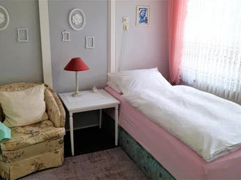 Ein einzelnes Bett mit weißer Bettwäsche und rosa Überwurf steht neben einem gepolsterten Sessel.