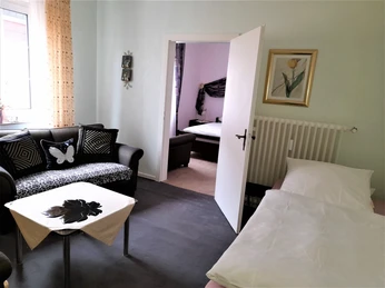 Helles Gästezimmer mit zwei Betten, Sofa und Tisch, dekoriert mit Blumenbild und Schmetterlingsmotiv.