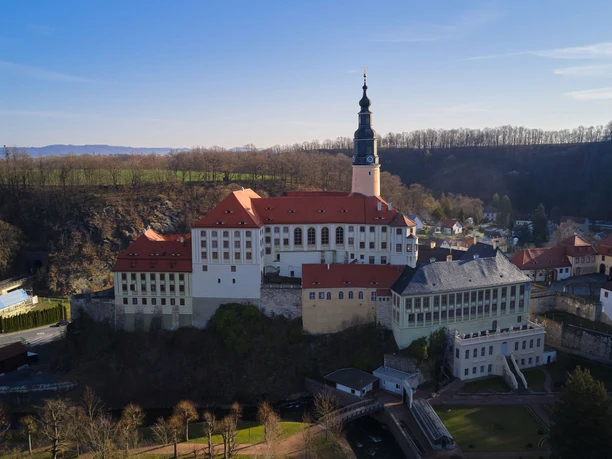 Schloss Weesenstein Luftaufnahme von Schloss Weesenstein mit roten Dächern und einem hohen Turm, umgeben von Bäumen und einer kleinen Siedlung bei klarem Himmel.