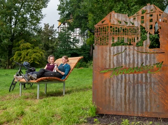 Zwei Frauen auf einer Landschaftsliege mit einer Cortenstahlarbeit der Tonenburg und der Tonnenburg im Hintergrund