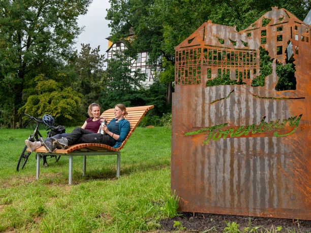 rastplatz_tonenburg_Henning_Orendt (8).jpg Zwei Frauen auf einer Landschaftsliege mit einer Cortenstahlarbeit der Tonenburg und der Tonnenburg im Hintergrund