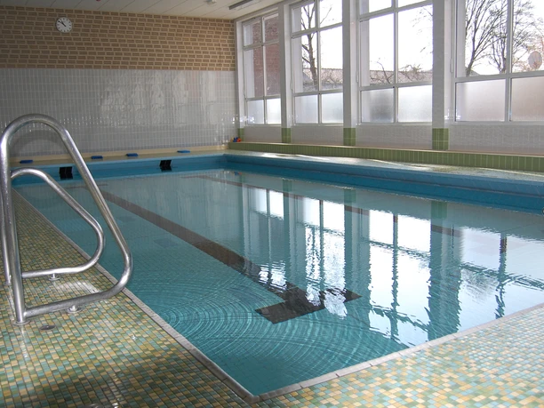 Schwimmhalle mit türkisfarbenem Becken, Stufen- und Railing-Zugang, großen Fenstern und heller Kachelwand.