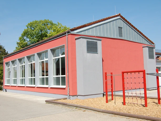Hallenbad Clarholz.JPG Rotes, modernes Gebäude mit großen Fenstern, Flachdach, neben hellem Spielplatz mit Klettergerüst.