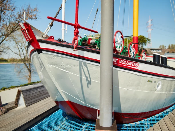 Aalfischerei-Museum in Monheim am Rhein Ein traditionelles Fischerboot namens "Voluntas" liegt auf einem erhöhten, modernen Netzdisplay im Freien.