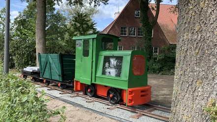 Lorenbahn.jpg Eine kleine grüne Lorenbahn mit roten Akzenten steht auf Schienen vor einem Backsteinhaus im Grünen.