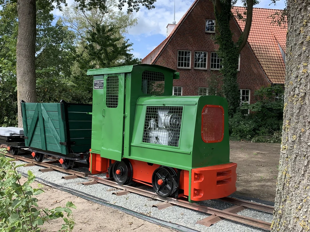 Lorenbahn.jpg Eine kleine grüne Lorenbahn mit roten Akzenten steht auf Schienen vor einem Backsteinhaus im Grünen.