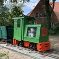 Lorenbahn.jpg Eine kleine grüne Lorenbahn mit roten Akzenten steht auf Schienen vor einem Backsteinhaus im Grünen.