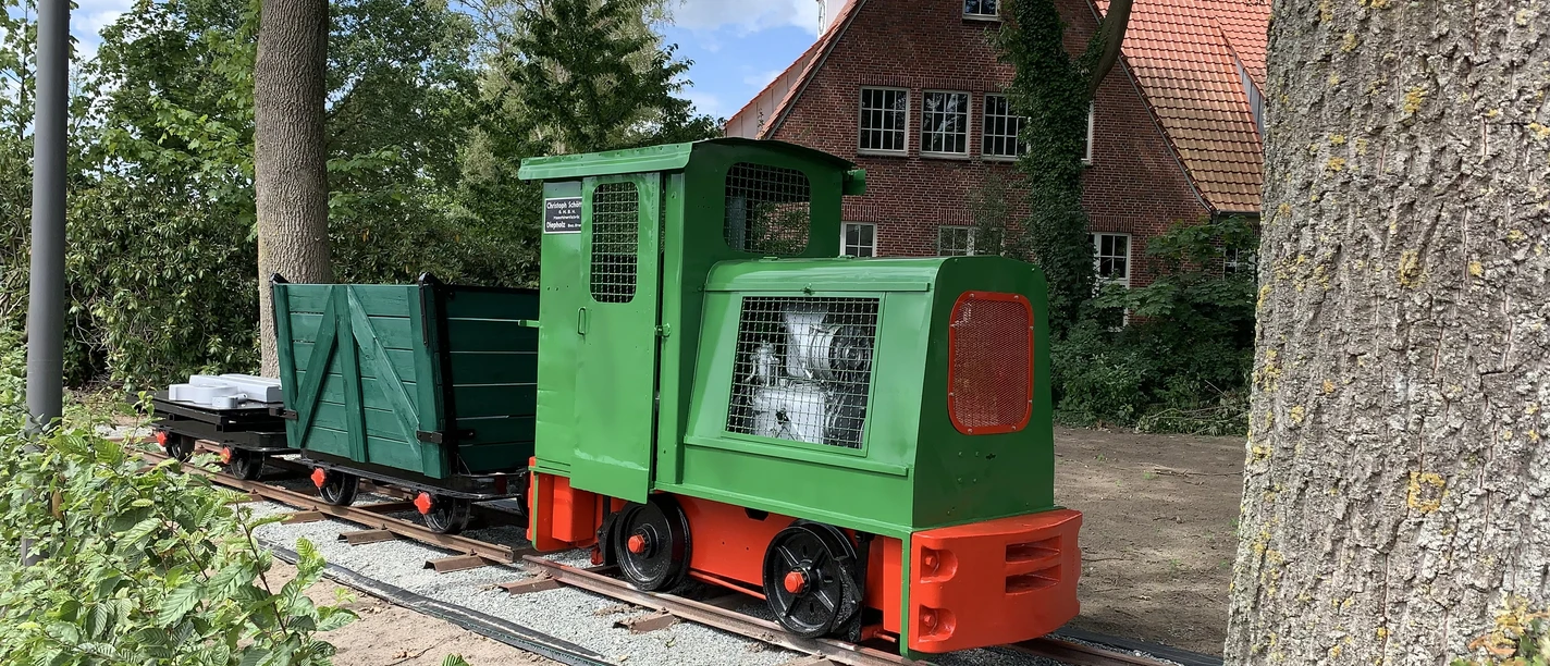 Lorenbahn.jpg Eine kleine grüne Lorenbahn mit roten Akzenten steht auf Schienen vor einem Backsteinhaus im Grünen.