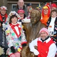 Karnevalistisch kostümierte Personen versammeln sich fröhlich um eine Bronzestatue bei einem Event im Freien.
