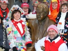 Karnevalistisch kostümierte Personen versammeln sich fröhlich um eine Bronzestatue bei einem Event im Freien.