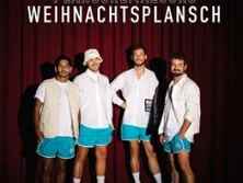 Vier Männer in blauen Shorts vor einem roten Vorhang, lächeln in die Kamera. Sie tragen Hemden.