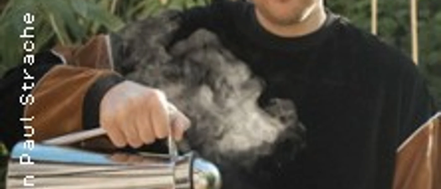 Person mit schulterlangem Haar gießt draußen ein dampfendes Getränk aus einer Kanne ein.