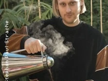 Person mit schulterlangem Haar gießt draußen ein dampfendes Getränk aus einer Kanne ein.