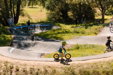 Kinder fahren mit Fahrrädern auf einem hügeligen Pumptrack umgeben von Bäumen und Wiesen.