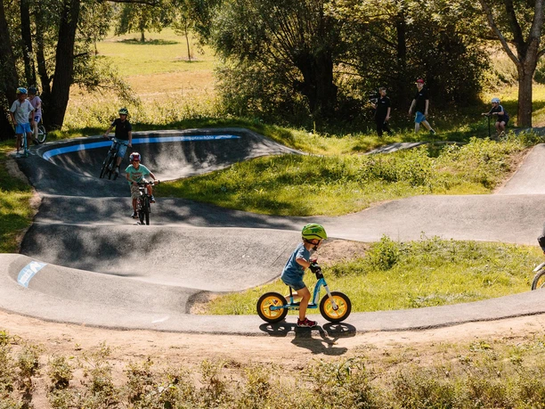 Kinder fahren mit Fahrrädern auf einem hügeligen Pumptrack umgeben von Bäumen und Wiesen.