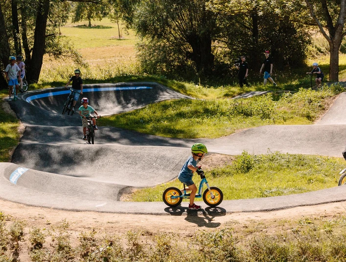 Kinder fahren mit Fahrrädern auf einem hügeligen Pumptrack umgeben von Bäumen und Wiesen.