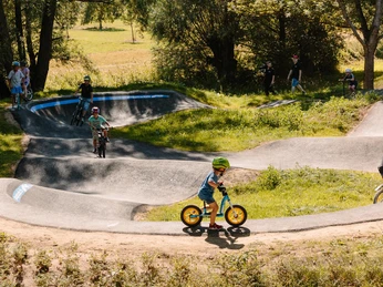 Pumptrack 4 - Kind mit Laufrad.jpg Kinder fahren mit Fahrrädern auf einem hügeligen Pumptrack umgeben von Bäumen und Wiesen.