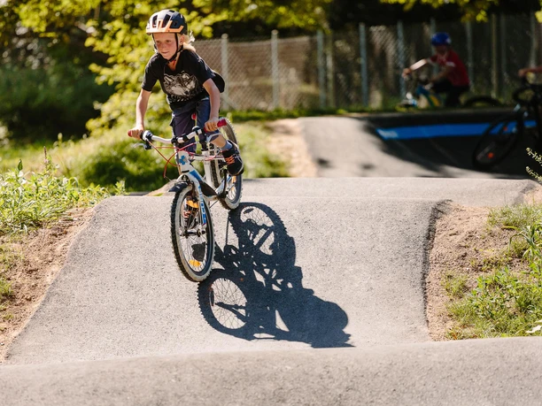 Ein Kind mit Helm fährt fröhlich auf einem BMX-Rad über eine asphaltierte Buckelpiste im Park.