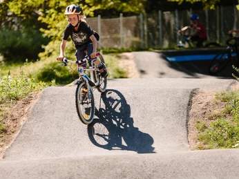 Pumptrack 1 - Kind auf Rad.jpg Ein Kind mit Helm fährt fröhlich auf einem BMX-Rad über eine asphaltierte Buckelpiste im Park.