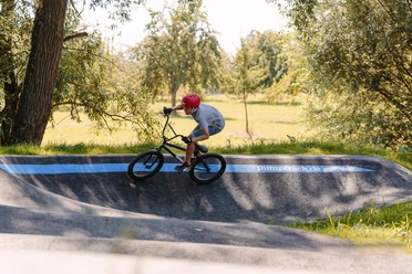 Ein Junge mit rotem Helm fährt auf einem BMX-Rad eine kurvige Asphaltstrecke entlang.