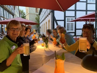 Gruppe älterer Menschen sitzt unter Sonnenschirmen an langen Tischen und genießt bunte Getränke.