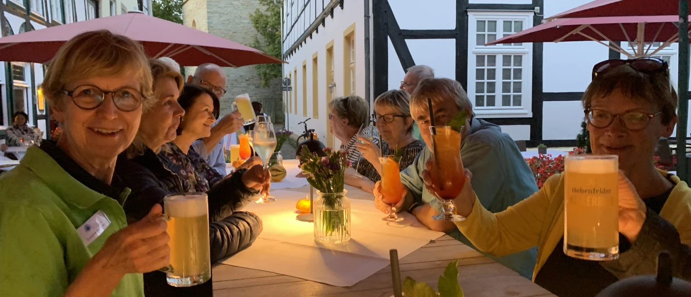 Gruppe älterer Menschen sitzt unter Sonnenschirmen an langen Tischen und genießt bunte Getränke.