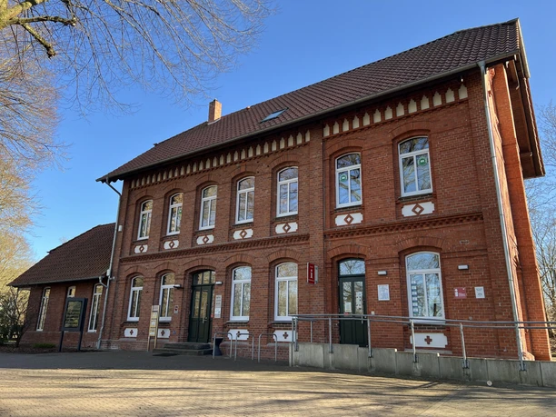 Backsteinbahnhof mit stufenlosem Zugang, gelb-braunes Dach, vor blauem Himmel und seitlichen Bäumen.