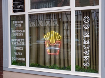 SnackNGo Auf dem Fenster steht das Angebot des Ladens: "French Fries, American Sweets, Mystery Packages, Drinks, Vapes, Popcorn"