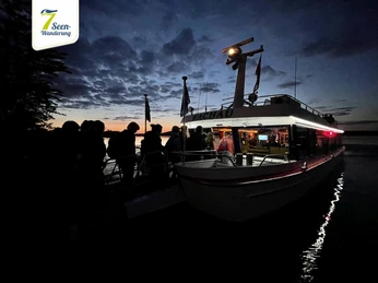 7-Seen-Wanderung©Sportfreunde_Neuseenland (4).jpg Menschen steigen bei Sonnenuntergang auf ein beleuchtetes Ausflugsschiff.People board an illuminated excursion boat at sunset.Lidé nastupují na osvětlenou výletní loď při západu slunce.Ludzie wsiadają na oświetloną łódź wycieczkową o zachodzie słońca.Mensen gaan aan boord van een verlichte excursieboot bij zonsondergang.Persone a bordo di una barca da escursione illuminata al tramonto.