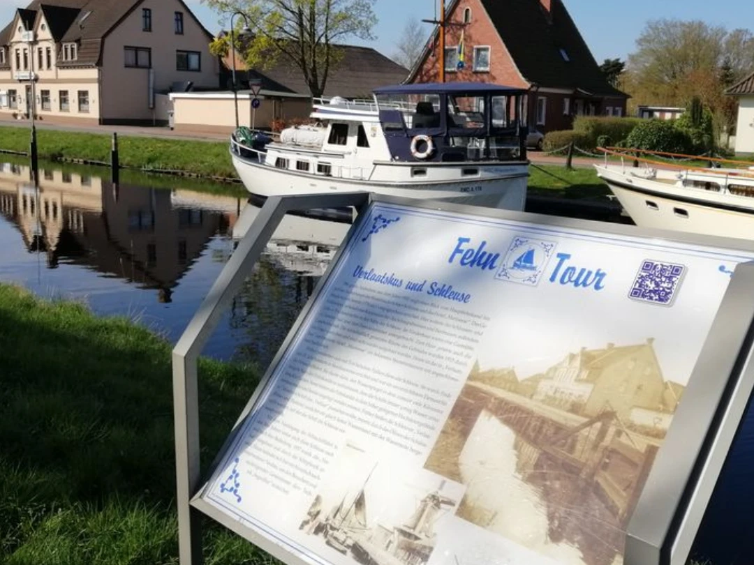 Fehn-Tour.png Infotafel zum historischen Rundgang vor einem Kanal mit Boot und Häusern in Rhauderfehn